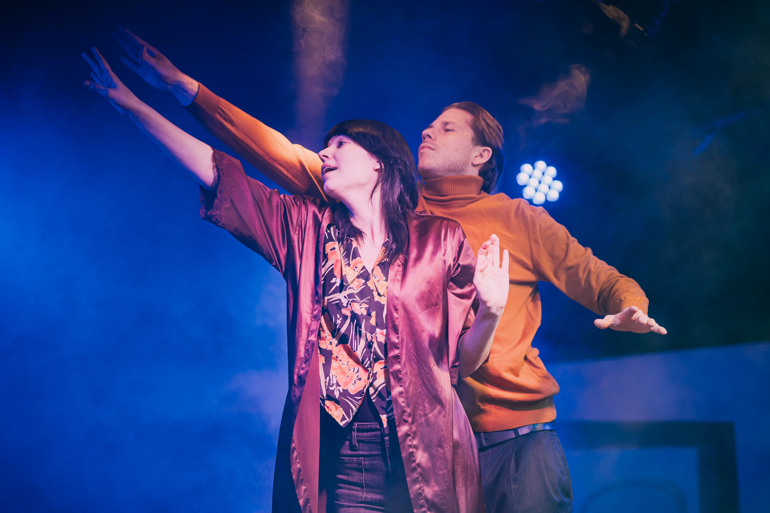 Na scenie teatralnej dwoje aktorów stoi blisko siebie w niebieskim świetle i lekkiej scenicznej mgle. Kobieta na pierwszym planie wyciąga rękę w górę i patrzy w tym samym kierunku, jakby wskazywała coś w oddali. Ma na sobie długi, satynowy szlafrok w odcieniu bordo i wzorzystą koszulę. Za nią stoi mężczyzna w pomarańczowym golfie, który również wyciąga rękę w tę samą stronę. Oboje przyjmują dynamiczną, niemal taneczną pozę, a w tle widoczne są sceniczne reflektory